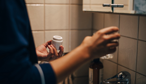 Överkropp och armar på en person som står i ett badrum med en burk medicin i ena handen och ett glas vatten i den andra.
